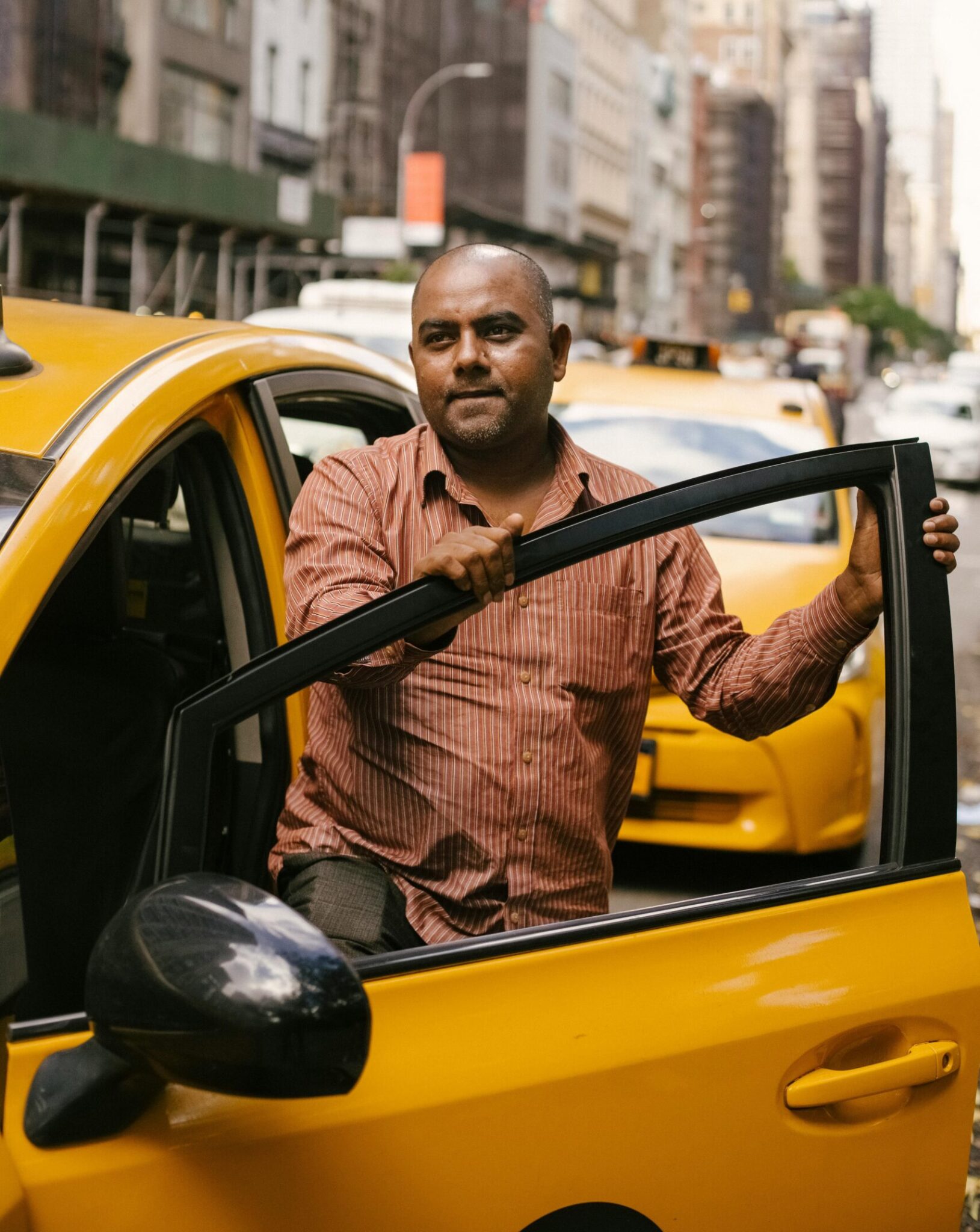 A taxi driver stepping out of a yellow cab in a busy city street during the day.